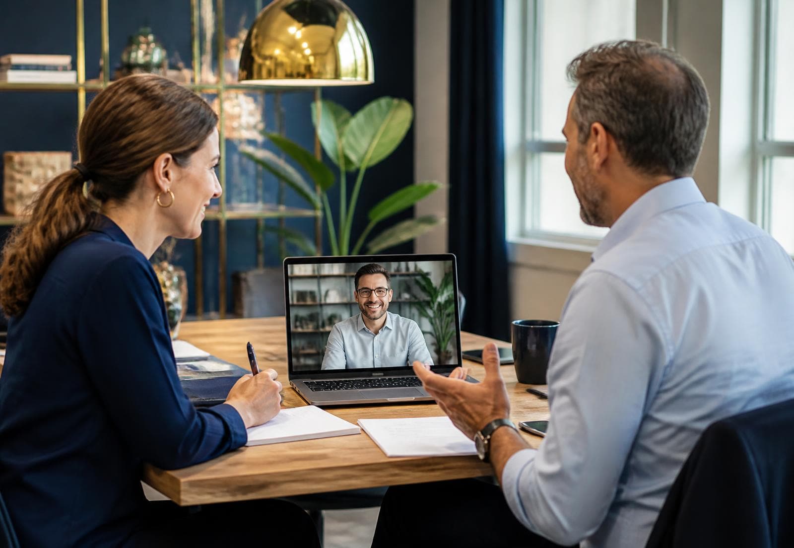 Two colleagues in an office on a video call with a remote teammate on a laptop